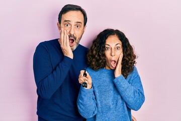 Middle age couple of hispanic woman and man holding keys of new home afraid and shocked, surprise and amazed expression with hands on face