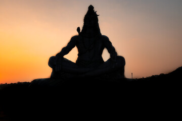 back lit statue of hindu god lord shiva in meditation posture with dramatic sky from unique angle