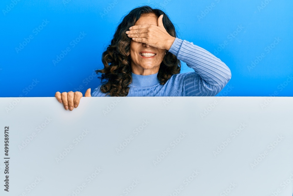 Wall mural middle age hispanic woman holding blank empty banner smiling and laughing with hand on face covering