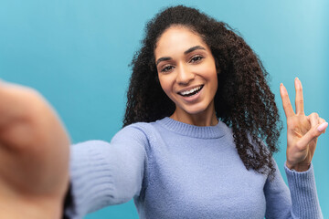 Cheerful young african-american woman in lavender sweater taking selfie, beautiful multiracial female student holding hand in victory gesture, smiling and looks at the camera isolated on pastel blue