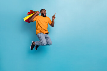 Obraz premium Photo of impressed funny guy dressed orange t-shirt jumping holding bargains pointing empty space isolated blue color background