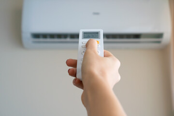 Close-up of a woman's hand directs the remote control to a white air conditioner on the wall, copy space