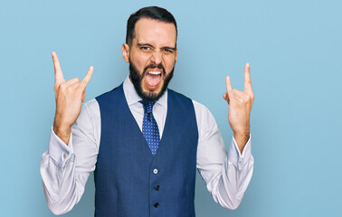 Young man with beard wearing business vest shouting with crazy expression doing rock symbol with hands up. music star. heavy concept.