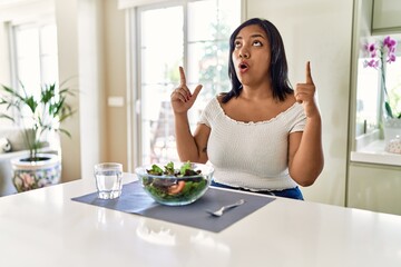 Obraz na płótnie Canvas Young hispanic woman eating healthy salad at home amazed and surprised looking up and pointing with fingers and raised arms.