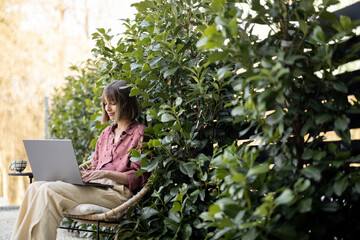 Young woman works on laptop computer while sitting relaxed on chair on background of green bushes...
