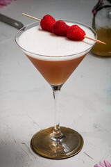 A French Martini cocktail garnished with fresh raspberries on a white background and styled with bar equipment and flowers.