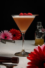 A French Martini cocktail garnished with fresh raspberries on a white background and styled with bar equipment and flowers.