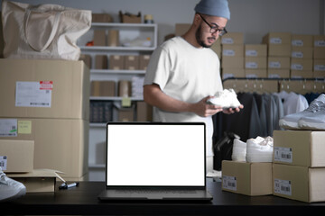 Laptop with blank white screen standing on the desktop in the warehouses. Asian man online seller checking stock and Inventory before sending to the customer. E-commerce business owner.