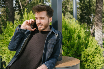 Young smiling man talking to girlfriend on phone. He is happy to talking with his girlfriend or friends. He is leaning against a table outdoors.