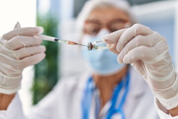 Senior grey-haired woman wearing doctor uniform and medical mask holding vaccine dose at clinic