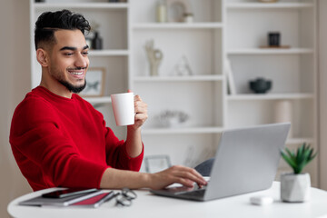 Cheerful attractive young arab businessman with beard in red clothes drinks coffee and types on...