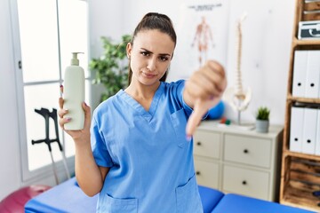 Young physiotherapist woman holding massage body lotion with angry face, negative sign showing...
