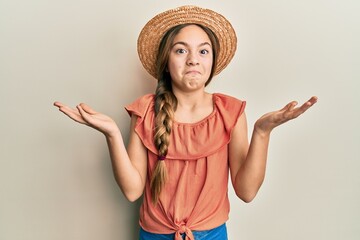 Beautiful brunette little girl wearing summer hat clueless and confused expression with arms and hands raised. doubt concept.