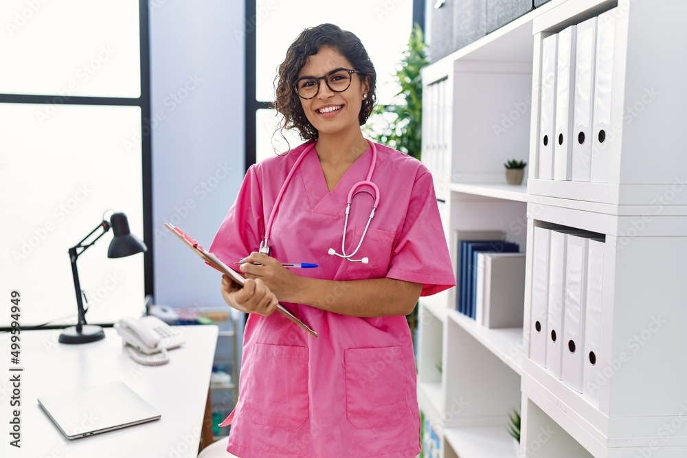 Wall mural young latin woman wearing doctor uniform writing on clipboard at clinic - Wall murals