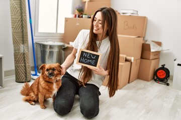 Young hispanic woman holding new home blackboard sitting on floor with dog at new home