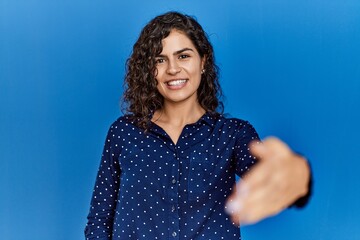 Young brunette woman with curly hair wearing casual clothes over blue background smiling friendly offering handshake as greeting and welcoming. successful business.