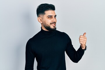 Fototapeta premium Handsome man with beard wearing turtleneck sweater pointing thumb up to the side smiling happy with open mouth