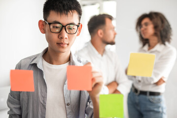 Focus ethnic guy taking notes on memo stickers during brainstorm with coworkers