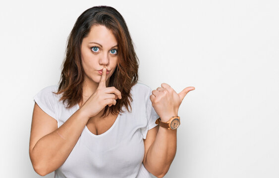 Young plus size woman wearing casual white t shirt asking to be quiet with finger on lips pointing with hand to the side. silence and secret concept.