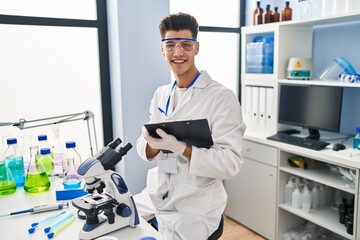 Naklejka na ściany i meble Young hispanic man wearing scientist uniform writing on clipboard at laboratory