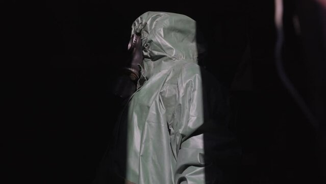 person dressed in protective anti chemical suit and in gas mask stands half dark and looks around closeup