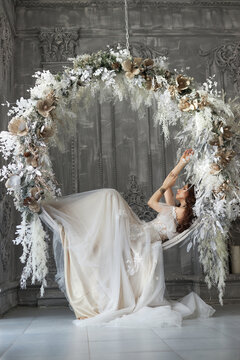 Woman Bride in a white dress swings on a round swing made of white flowers and feathers. A romantic image of a woman with a tattoo on her arm. Wedding