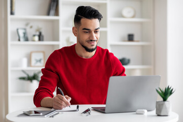 Smiling calm confident attractive millennial arab guy with beard in red clothes makes notes, works...