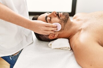 Fototapeta na wymiar Young hispanic man having clean facial treatment using sponge at beauty center