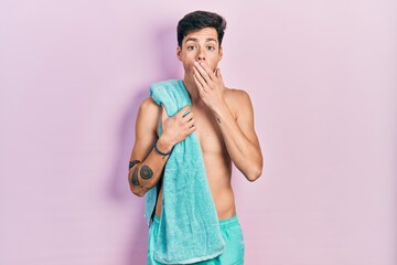 Young hispanic man wearing swimwear and towel covering mouth with hand, shocked and afraid for...