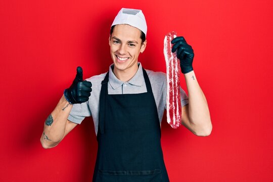 Young hispanic man wearing cook apron and holding meat smiling happy and positive, thumb up doing excellent and approval sign