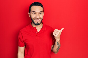 Fototapeta premium Hispanic man with beard wearing casual red t shirt smiling with happy face looking and pointing to the side with thumb up.