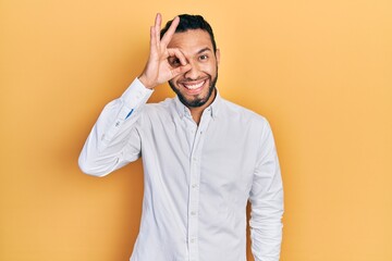 Hispanic man with beard wearing business shirt doing ok gesture with hand smiling, eye looking through fingers with happy face.