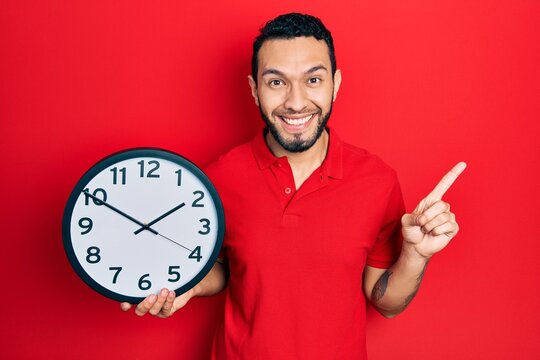 Hispanic man with beard holding big clock smiling happy pointing with hand and finger to the side