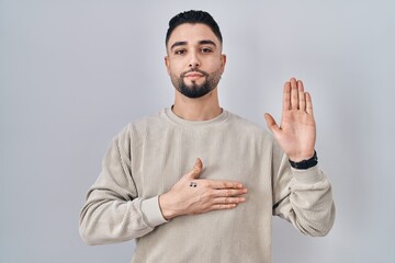 Young handsome man standing over isolated background swearing with hand on chest and open palm, making a loyalty promise oath