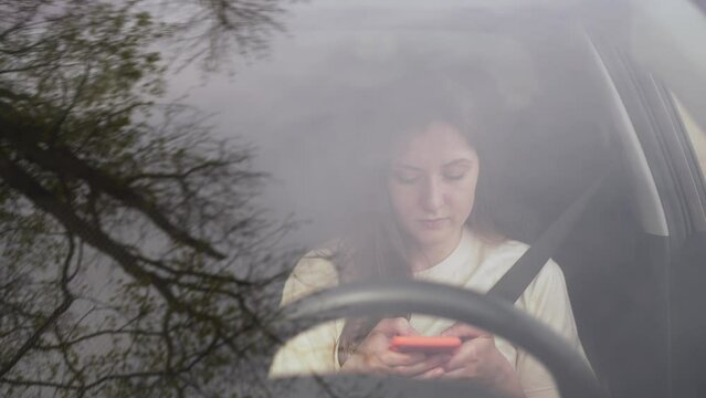 Portrait of a young woman texting on her phone while driving. Inattentive and distracted dangerous driver. Person writes a message inside the car standing in a traffic jam