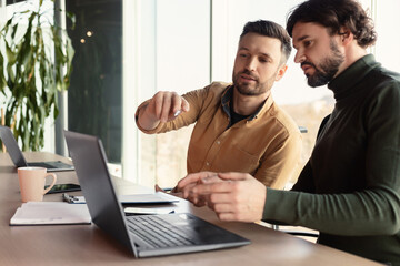 Obraz premium Serious employee speaking to his male colleague at company office, using laptop pc, discussing common business project