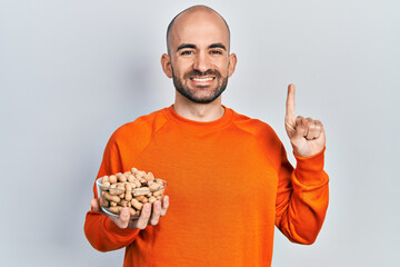 Young bald man holding peanuts smiling with an idea or question pointing finger with happy face, number one