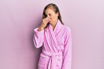 Young blonde woman wearing bathrobe smelling something stinky and disgusting, intolerable smell,...