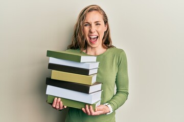 Young blonde woman holding a pile of books smiling and laughing hard out loud because funny crazy joke.