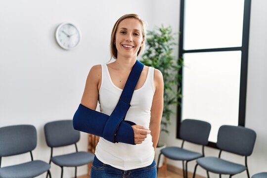 Young caucasian woman wearing sling on arm for accident standing at clinic waiting room