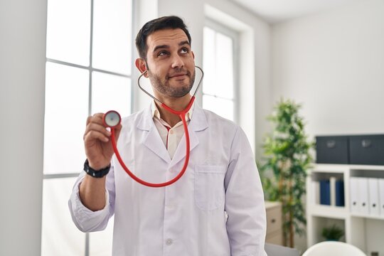 Young hispanic doctor man with beard holding stethoscope auscultating smiling looking to the side and staring away thinking.