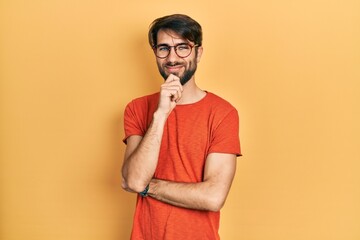 Young hispanic man wearing casual clothes and glasses looking confident at the camera with smile with crossed arms and hand raised on chin. thinking positive.