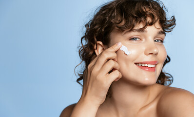 Close up portrait of smiling young woman with curly hair, using facial skincare cream on her face, doing cosmetic daily routine after shower, standing over blue background