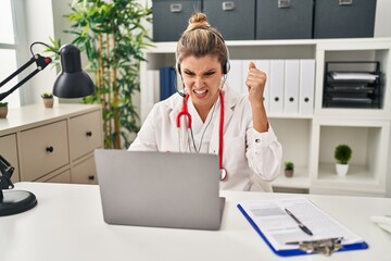 Young doctor woman wearing doctor uniform working using computer laptop annoyed and frustrated shouting with anger, yelling crazy with anger and hand raised