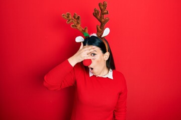 Young hispanic woman wearing deer christmas hat and red nose peeking in shock covering face and eyes with hand, looking through fingers with embarrassed expression.