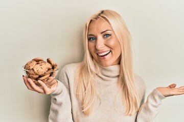 Young blonde woman holding bowl with chocolate chips cookies celebrating achievement with happy...