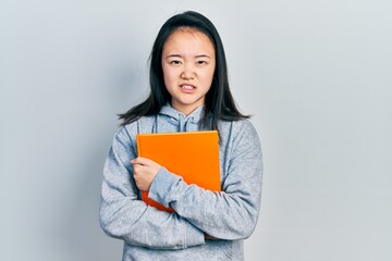 Obraz premium Young chinese girl holding book clueless and confused expression. doubt concept.