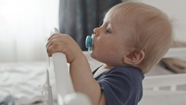 Side view of blond-haired Caucasian toddler with pacifier in mouth standing in crib at daytime in nursery