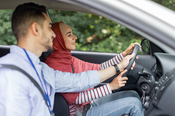 Happy attractive millennial arabic man teaching student, helping to woman in hijab at steering wheel to driving car