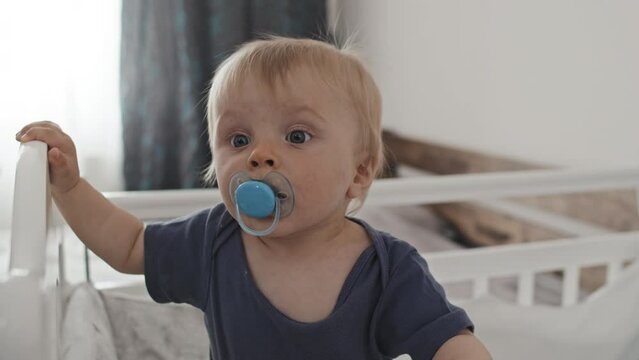 Waist-up of blond-haired Caucasian toddler with pacifier in mouth standing in crib at daytime, looking on camera